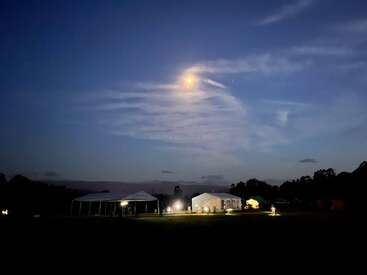 A serene night scene shows illuminated tents on a grassy field beneath a glowing moon and wispy clouds, with trees silhouetted in the background and calm atmosphere.