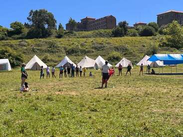 A group of people, mostly children, stands in a circle on a grassy field with tents behind them, participating in outdoor activities under a clear blue sky.