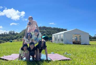 Six girls form a human pyramid outdoors on yoga mats, enjoying a sunny day with a large white tent and green hills in the background.
