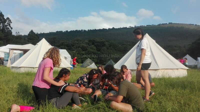 The image depicts a group of people sitting in a grassy field, surrounded by tents, with a serene forested hill and blue sky with clouds in the background.