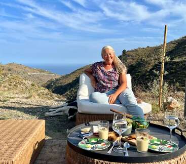 Das Bild zeigt eine ruhige Umgebung im Freien mit einer Frau, die auf einer weißen Couch sitzt, umgeben von einem Tisch mit Essen und Getränken, vor einer malerischen Kulisse aus Hügeln und blauem Himmel.