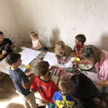 Mehrere Kinder und zwei Erwachsene sind um einen Tisch versammelt und basteln mit Buntstiften, Farben und Naturmaterialien in einem gemütlichen Raum.