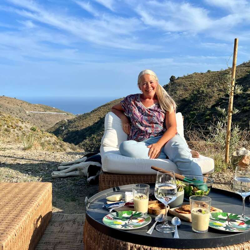 Das Bild zeigt eine ruhige Umgebung im Freien mit einer Frau, die auf einer weißen Couch sitzt, umgeben von einem Tisch mit Essen und Getränken, vor einer malerischen Kulisse aus Hügeln und blauem Himmel.