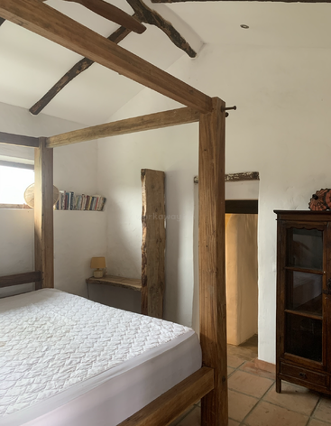 L'image représente une pièce avec un lit, une table et une chaise, aux murs et au sol blancs, avec une grande fenêtre sur le côté gauche de l'image.