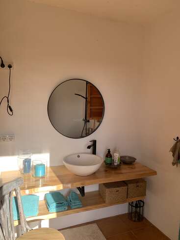 A minimalist bathroom features a round mirror, vessel sink, light wooden shelves, blue towels, candles, baskets for storage, and natural sunlight illuminating the cozy space.