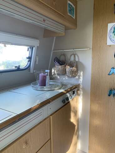 This image shows a cozy camper kitchen with wooden cabinets, a window, a small stovetop, hanging baskets, decorative candles, and sunlight casting soft shadows.