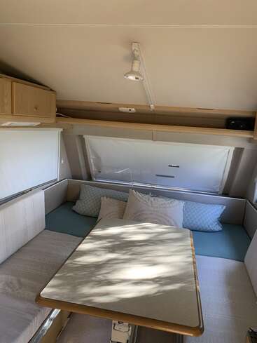 The image shows a cozy caravan interior with a table, surrounding cushioned bench seats, pillows, overhead cupboard, large window shade, and a ceiling light. Sunlight filters in.