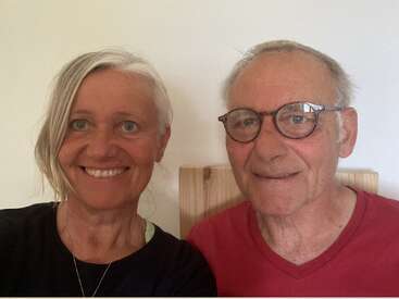 A woman with short, light hair and a man with glasses smile for a selfie, sitting close together indoors against a plain, light-colored wall.