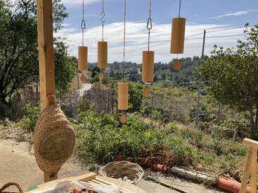 Wooden wind chimes hang from strings on a porch overlooking a lush green garden and distant hills, with a woven hat and baskets on the table.
