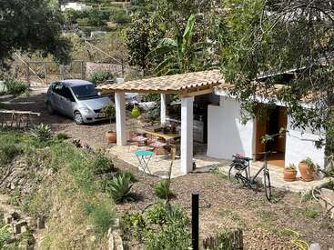 A small white house with a tiled roof stands surrounded by plants. There’s a parked car, bicycle, outdoor dining table, greenery, and a peaceful rural atmosphere.