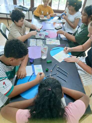 Un grupo de niños sentados alrededor de una mesa dibujan, colorean y hacen manualidades con rotuladores, lápices y papel, concentrados en su creativo proyecto escolar.