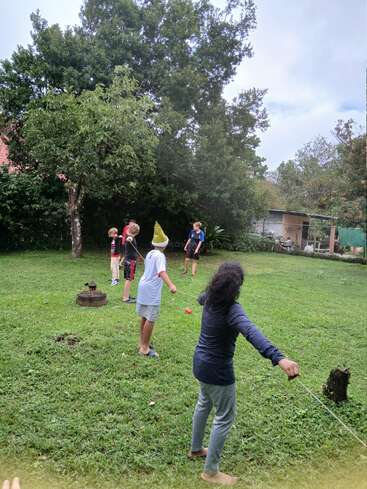 Cinco niños juegan en un prado rodeados de árboles y vegetación. Uno de los niños lleva un gorro en forma de cono amarillo. Parece divertido.