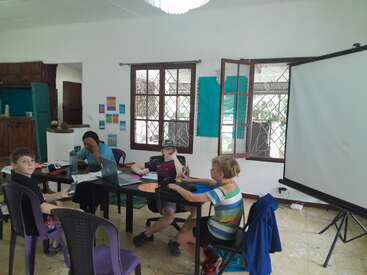 Vier Kinder sitzen um einen Tisch in einem hellen Klassenzimmer und arbeiten mit Laptops und Notebooks. Offene Fenster, bunte Wände und eine entspannte Atmosphäre lassen auf eine lockere, kreative Lernumgebung schließen.