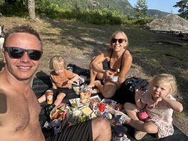 Uma família de quatro pessoas curte um piquenique ao ar livre. Eles estão sorrindo, vestindo roupas de verão e óculos escuros, rodeados de comida e bebida, no meio da natureza.