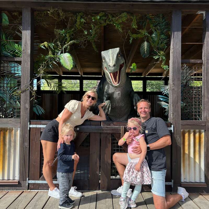 Uma família sorridente de quatro pessoas posa na frente de uma estátua de dinossauro super realista num recinto ao ar livre, cercada por plantas verdes e estruturas de madeira.