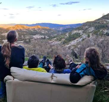 Vier Menschen entspannen sich auf einer Couch im Freien und genießen bei Sonnenuntergang den Blick auf ein malerisches Bergtal. Gemeinsam genießen sie die friedliche Aussicht, umgeben von Natur und Schönheit.