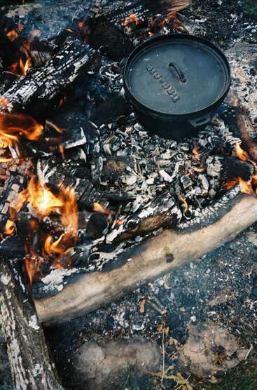 Una olla de hierro fundido negro descansa sobre las brasas incandescentes y los leños ardientes de una hoguera, rodeada de cenizas y fuego, al aire libre en un entorno rústico.