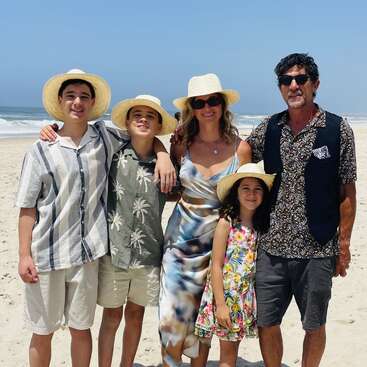 A family of five, wearing summer clothes and straw hats, stands smiling together on a sunny beach with blue sky and ocean waves in the background.