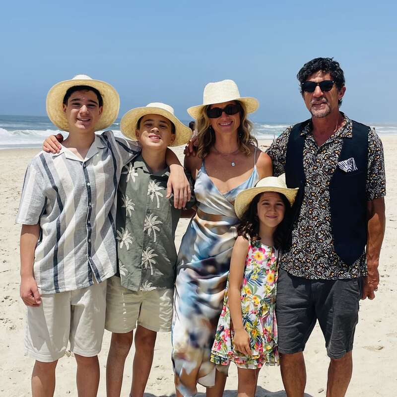 A family of five, wearing summer clothes and straw hats, stands smiling together on a sunny beach with blue sky and ocean waves in the background.