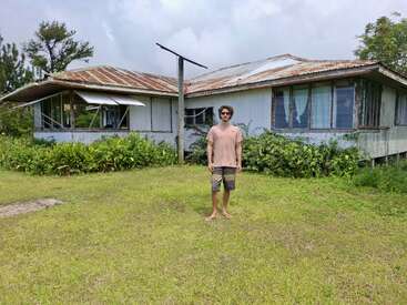 Par une journée nuageuse, un homme se tient pieds nus sur l'herbe devant une vieille maison usée par les intempéries, au toit de tôle rouillée et aux plantes envahissantes.