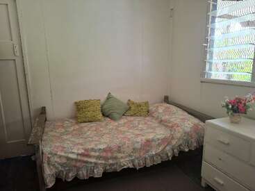 Cette chambre confortable comprend un lit de jour recouvert de fleurs avec trois oreillers décoratifs, une commode blanche avec des tiroirs, un vase de fleurs et une fenêtre avec des stores.