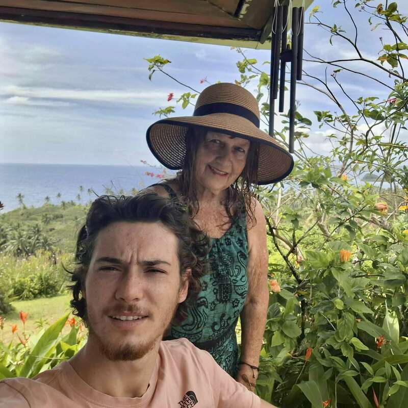 Um homem jovem e uma mulher mais velha, ambos sorridentes, posam ao ar livre cercados por vegetação exuberante, flores e vista para o mar. A mulher usa um chapéu de palha de abas largas.