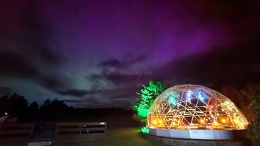 Eine leuchtende geodätische Kuppel, die von bunten Lichtern beleuchtet wird, steht nachts auf einer Wiese unter einem dramatischen Himmel, der in Lila- und Blautönen gemalt ist.