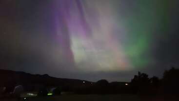 Dieses Bild zeigt die Nordlichter mit leuchtenden grünen und violetten Farben, die den Nachthimmel über einer silhouettenhaften Landschaft erhellen und eine magische, ätherische Atmosphäre über dem Horizont schaffen.