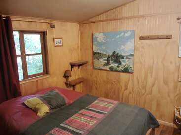 Esta acogedora habitación cuenta con paredes de madera, una cama doble con almohadas y sábanas de colores, una ventana con cortinas, un cuadro junto al mar y una sencilla decoración rústica.
