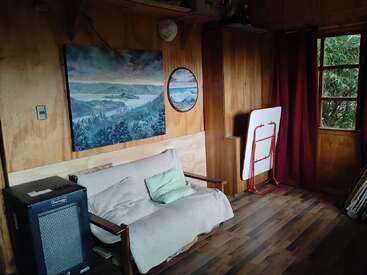 Esta acogedora habitación de madera cuenta con un pequeño sofá con cojines, pinturas murales, un calefactor, una mesa plegada, cortinas rojas y una ventana con vistas a la vegetación.
