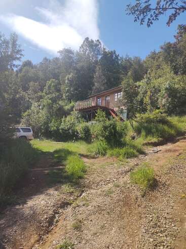 Una casa de madera sobre pilotes se alza en medio de una exuberante vegetación y árboles, con un coche blanco aparcado cerca. La luz del sol y el cielo azul crean un ambiente tranquilo.