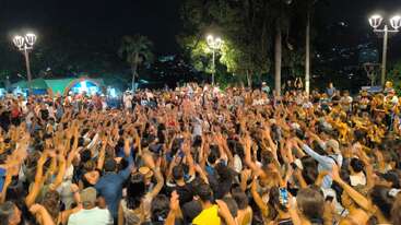 Uma galera grande e animada se junta ao ar livre à noite, levantando as mãos com entusiasmo. As pessoas estão envolvidas, iluminadas pelas luzes da rua, curtindo um evento comunitário animado sob as árvores.
