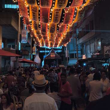 Um animado festival de rua à noite apresenta luzes e faixas vibrantes, uma grande multidão curtindo música, um ambiente alegre e barracas de comida alinhadas em ambos os lados.