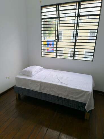 Um quarto simples com paredes brancas, piso de madeira, uma cama de solteiro coberta com lençóis brancos, um travesseiro e uma grande janela com grades de metal com vista para o exterior.