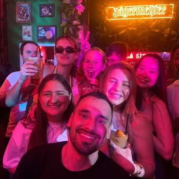Um grupo de amigos sorri e posa para uma foto em um bar animado, com bebidas nas mãos, sob luzes coloridas, com um letreiro da Jägermeister brilhando atrás deles.