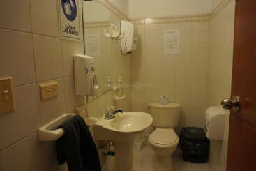 This image shows a small, clean public restroom with a sink, mirror, toilet, hand towel, soap dispenser, waste bin, and signs in Spanish.