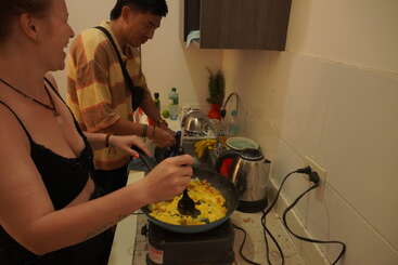 Two people cook together in a small kitchen, smiling and enjoying the process. Vegetables and eggs are being prepared in a pan on the stove.
