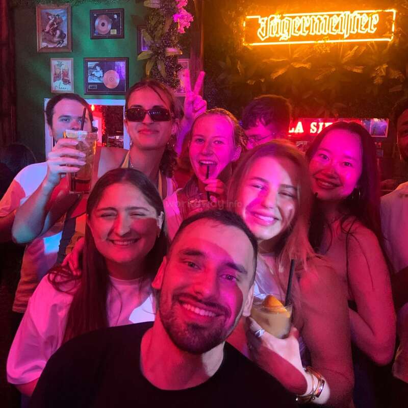 Um grupo de amigos sorri e posa para uma foto em um bar animado, com bebidas nas mãos, sob luzes coloridas, com um letreiro da Jägermeister brilhando atrás deles.