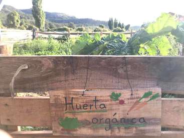 The image depicts a wooden sign with the words \"Huerta Organica\" in front of a garden, featuring a fence and trees in the background on a sunny day.