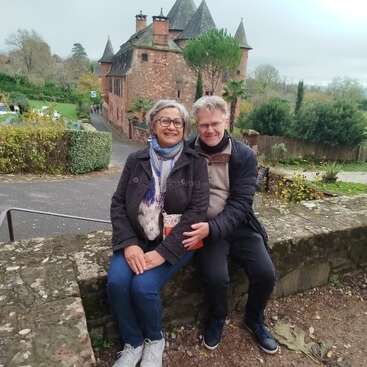 Um casal sorridente está sentado em um muro de pedra em frente a um castelo histórico de tijolos, cercado por vegetação e um ambiente tranquilo ao ar livre, aproveitando o dia.