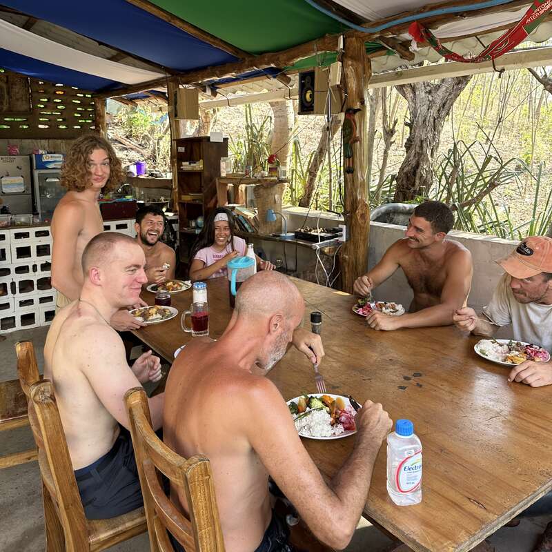 Sieben Leute sitzen an einem Holztisch unter einem bunten Sonnendach und essen zusammen. Die Stimmung ist locker und freundlich, und es wird viel gelacht und geredet.