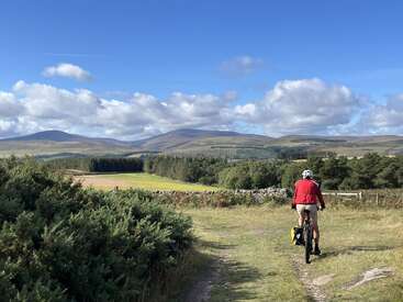 A cyclist in a red jacket rides along a rural dirt path, surrounded by green fields, hills, and trees under a bright blue sky with clouds.