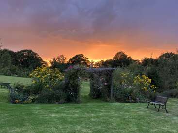 A beautiful garden at sunset features vibrant flowers, a wooden pergola, green grass, benches, and trees, all beneath a dramatic orange and purple sky.