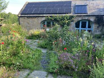 A charming stone cottage with solar panels sits behind a lush, colorful wildflower garden. Ivy climbs the facade, while vibrant blossoms line a stone path.