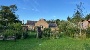 A quaint stone cottage sits amidst lush greenery and gardens under a clear blue sky. Benches and flourishing plants create a peaceful countryside atmosphere.