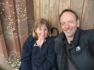 The image depicts a middle-aged couple posing for a selfie in front of a rustic wooden door and stone wall, both smiling at the camera.