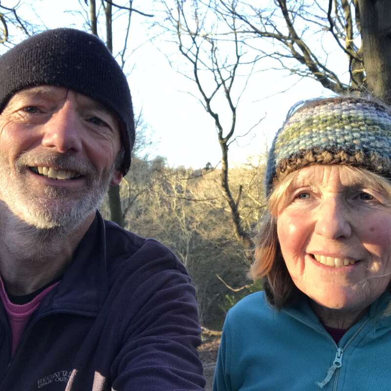 An older man and woman, both smiling, are outdoors on a sunny day. They wear warm hats and jackets, with bare trees visible in the background.
