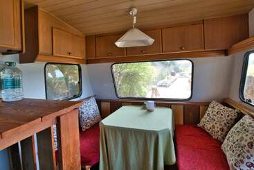 Cette image montre l'intérieur confortable d'un camping-car avec des armoires en bois, des banquettes rembourrées rouges, des coussins à motifs floraux, une nappe verte, de grandes fenêtres, une bouteille d'eau et une vue sur l'extérieur.