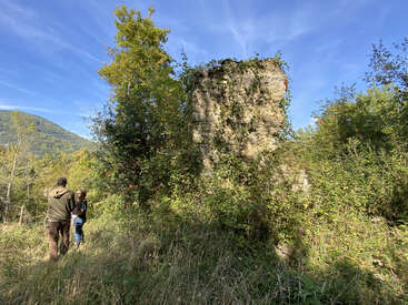 Deux personnes se tiennent dans une clairière herbeuse près de vieilles ruines de pierre envahies par la végétation. Une végétation luxuriante les entoure, avec des collines et un ciel bleu en arrière-plan, suggérant une exploration.