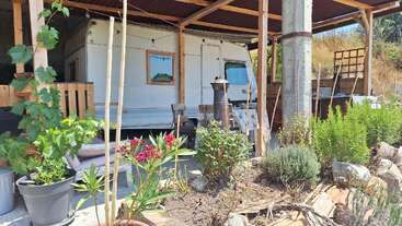 Un aménagement extérieur confortable comprend une caravane sous un toit en bois, entourée de plantes en pot luxuriantes, de parterres de fleurs, de chaises et d'une table, créant ainsi un havre de paix propice à la détente.
